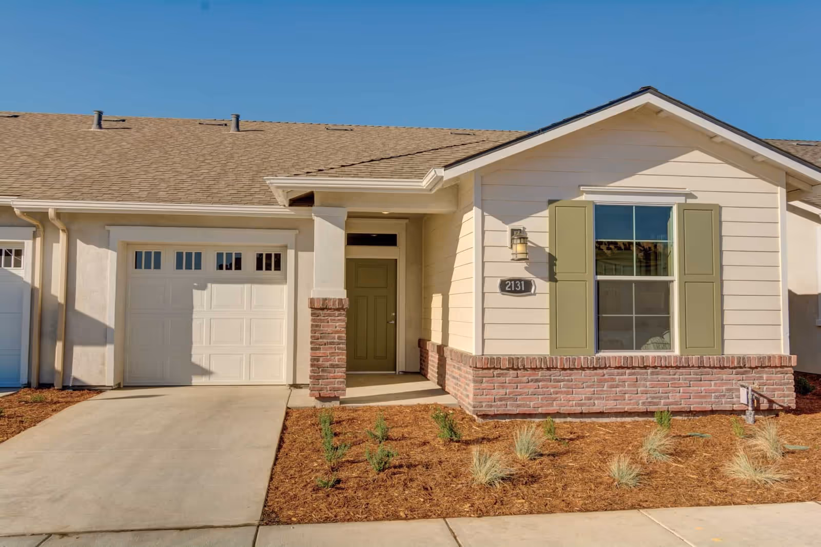 Exterior view of a single-story residential unit with a beige facade, green shutters, a green front door, a garage door, and a small landscaped area with young plants in front.