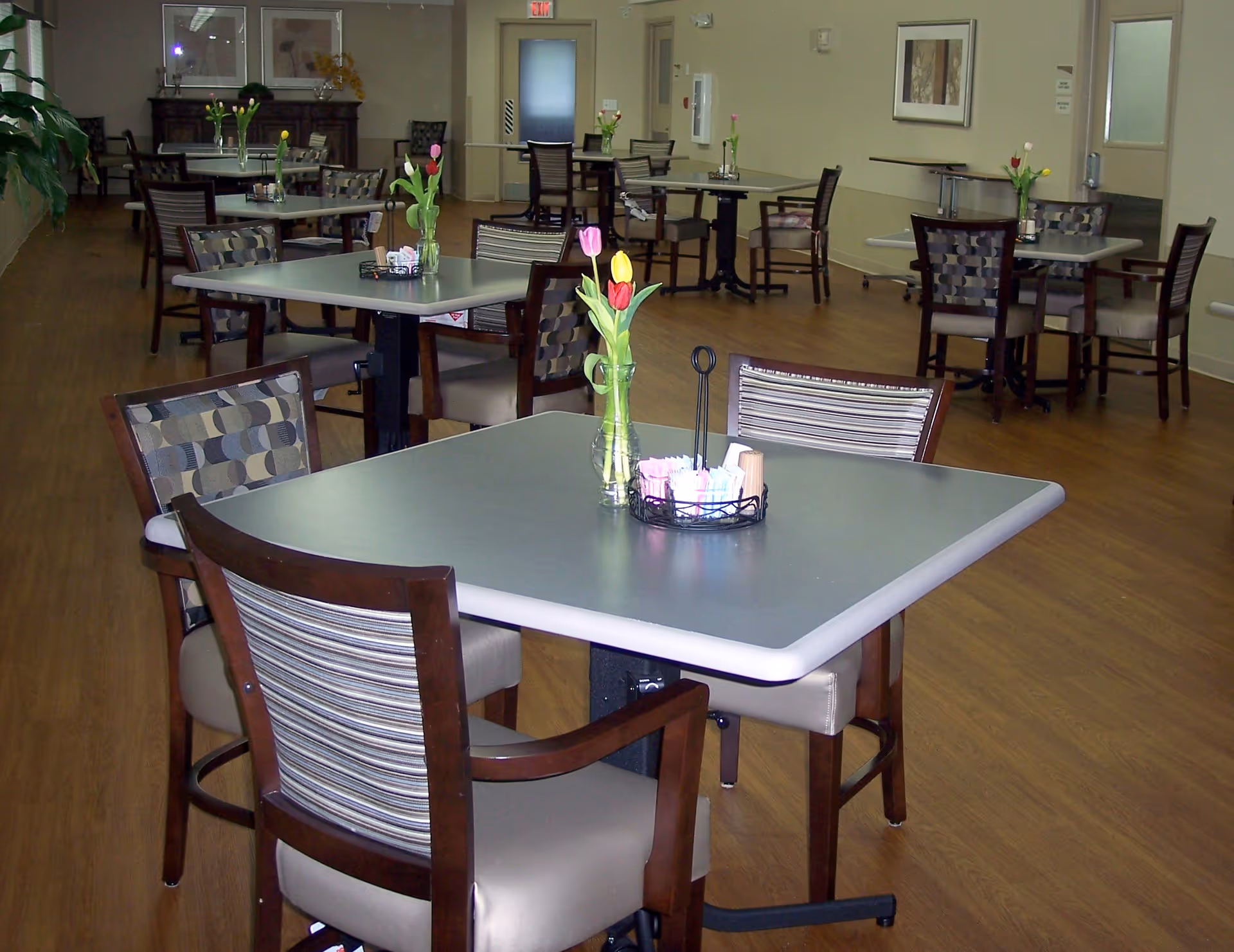 A dining room in a senior living facility with multiple square tables and chairs arranged neatly. Each table has a small vase with colorful tulips and a container holding sugar packets. The room has wood flooring, light-colored walls, framed artwork, and a door at the far end.
