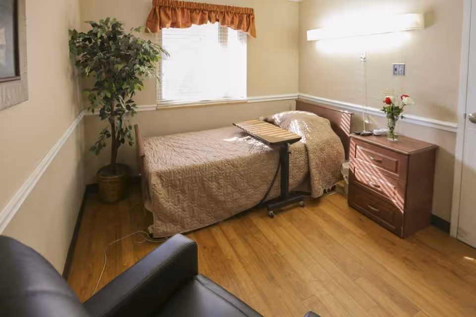Sunny single-occupancy bedroom with a bed, overbed table, wooden nightstand with flowers, potted plant, and an armchair.