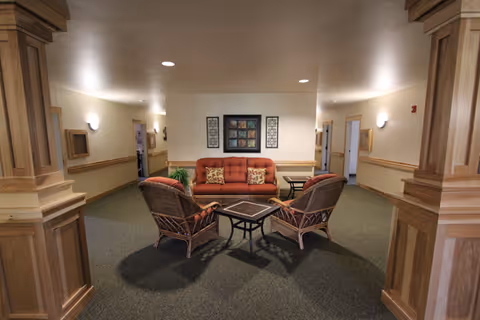 A cozy seating area in a hallway with a red sofa, two wicker chairs with cushions, and a small square table in the center. The walls are light-colored with wooden trim and decorative columns framing the space. There is a framed artwork on the wall behind the sofa and soft lighting from wall sconces.