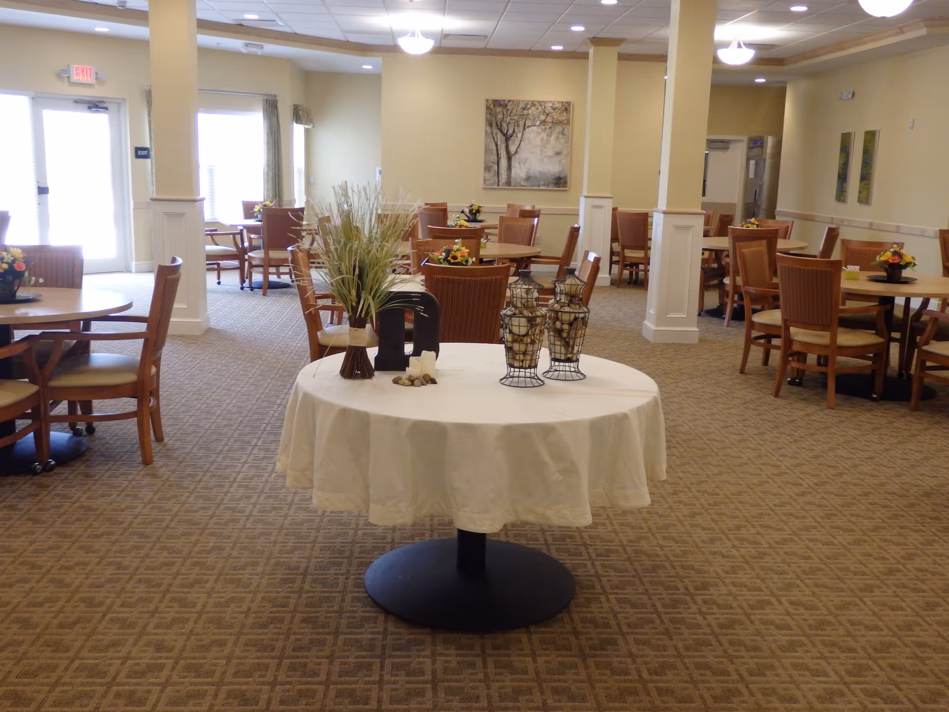 A spacious dining room with multiple round wooden tables and chairs arranged neatly. A round table in the center is covered with a white tablecloth and decorated with a vase of dried plants and two decorative wire baskets filled with white balls. The room has beige walls, carpeted floor with a geometric pattern, and several pieces of wall art. Large windows and an exit door are visible on the left side.