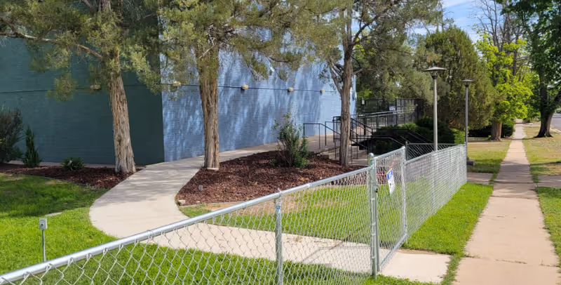 Outdoor view of a building with a light blue painted wall, a curved concrete walkway, trees, and a chain-link fence along a grassy area next to a sidewalk.