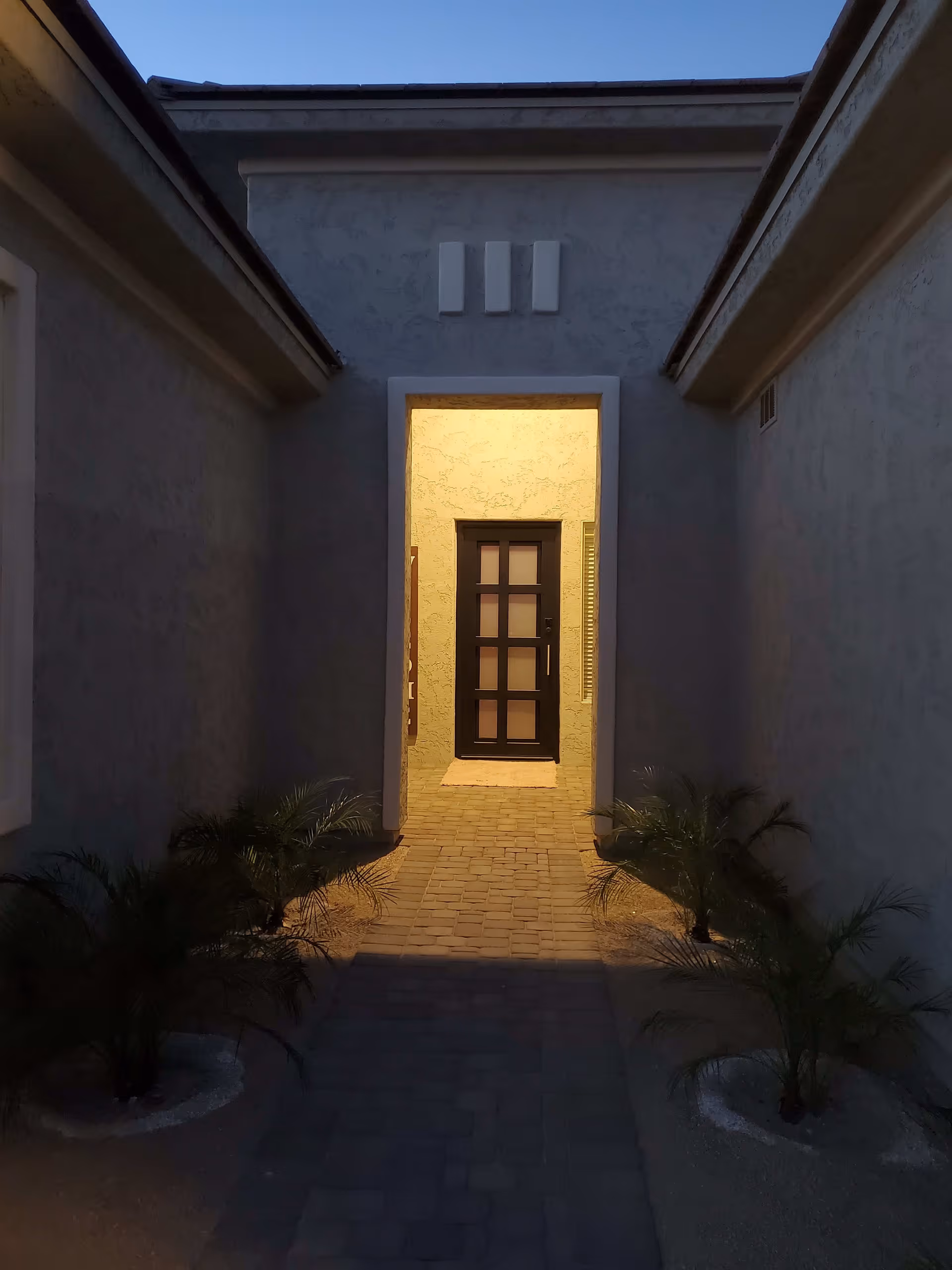 A dimly lit outdoor walkway leading to a modern black door with frosted glass panels. The walkway is paved with bricks and flanked by small palm plants on both sides. The walls on either side are light-colored stucco, and the sky above is clear and darkening, indicating dusk or early evening.