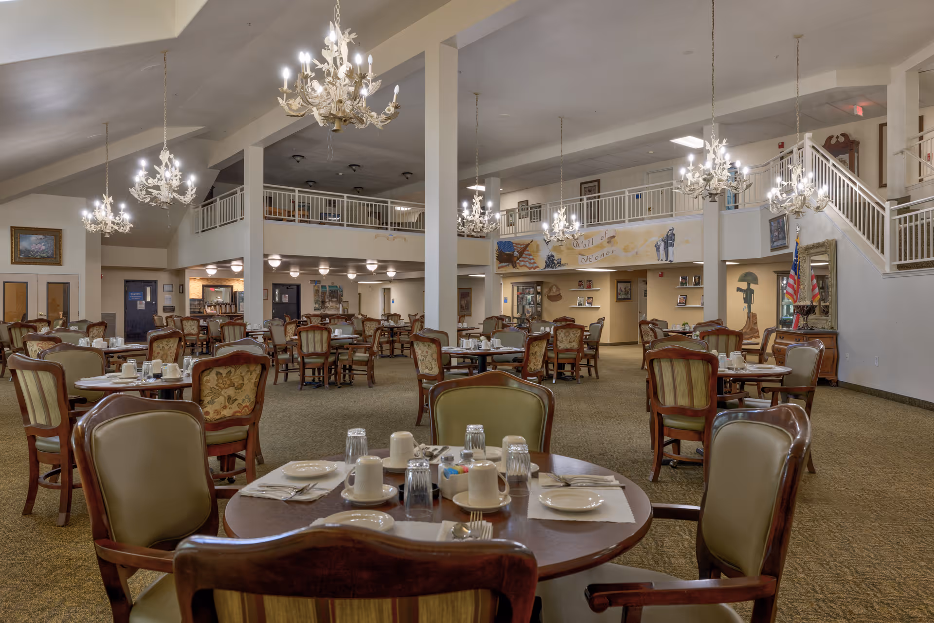 A spacious dining room in a senior living facility with multiple round tables set with plates, cups, and utensils. The room features elegant chandeliers hanging from a high ceiling, upholstered chairs, and a carpeted floor. There is a mezzanine level with railings and a staircase on the right side. The walls are decorated with paintings and a mural.
