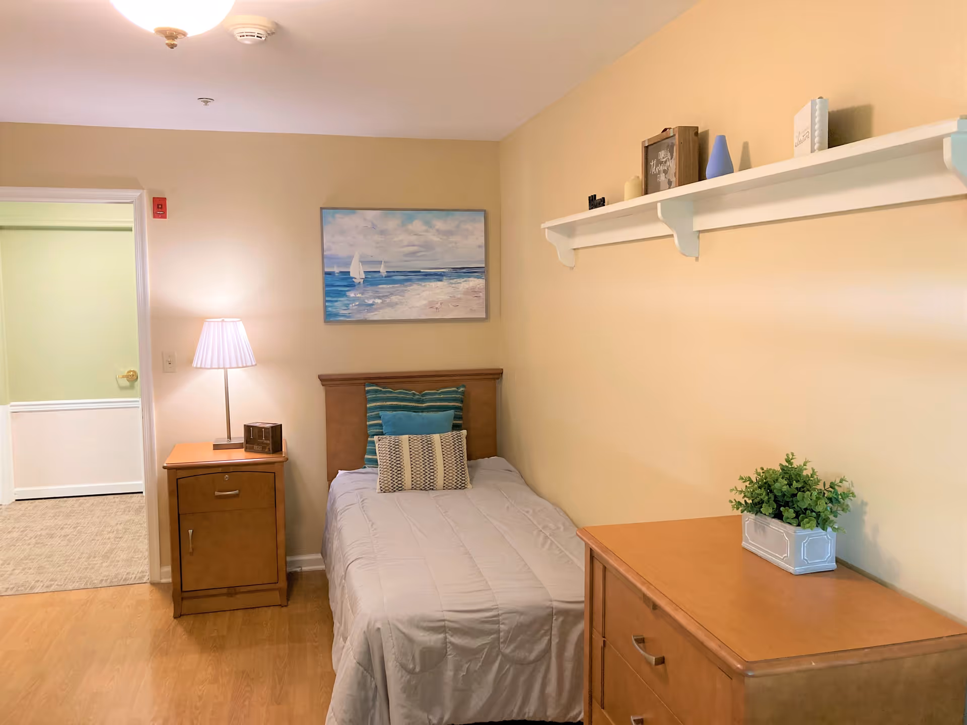 A small bedroom with a single bed against a beige wall. The bed has a gray comforter and three decorative pillows. Next to the bed is a wooden nightstand with a lamp and a small clock. Above the bed is a painting of sailboats on the ocean. On the right side of the room is a wooden dresser with a small potted plant on top. A white shelf with decorative items is mounted on the wall above the dresser. The floor is wooden, and an open doorway leads to another room with carpeted flooring.