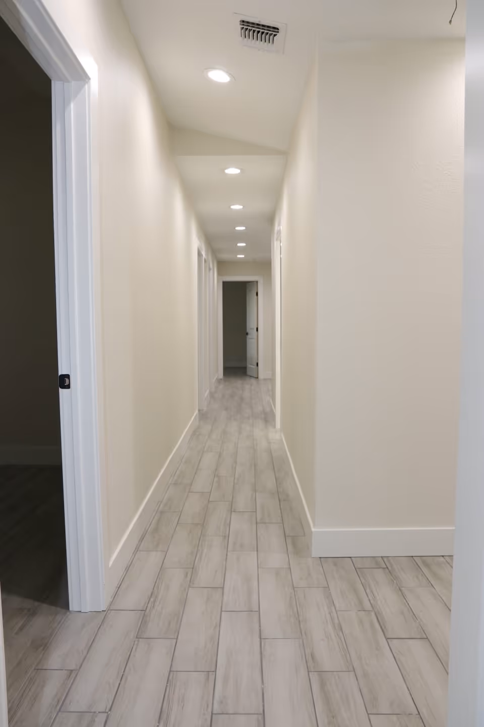 A long, narrow hallway with light-colored walls and wood-look tile flooring. Several recessed ceiling lights illuminate the corridor, and multiple doorways are visible along the hallway, with one door open at the far end.