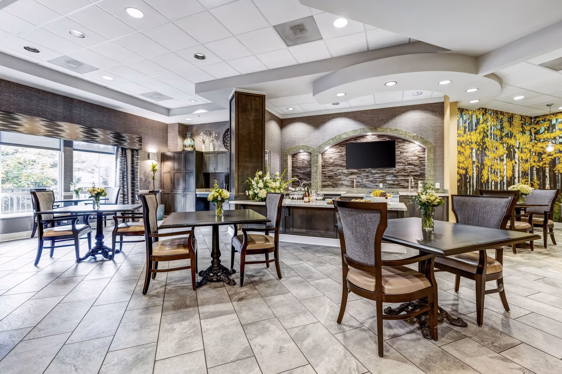 A bright and spacious dining area with several dark wood tables and chairs, each table decorated with a vase of flowers. The room features large windows with patterned curtains, a tiled floor, and a modern kitchen area in the background with a TV mounted on a stone accent wall. A colorful mural of yellow trees decorates one wall.