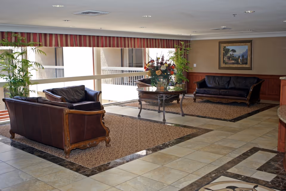 A spacious and well-lit common area with two dark brown leather sofas and a matching armchair arranged around a round glass-top table with a large floral arrangement. The floor is tiled with decorative rugs under the seating areas. There are large windows with striped valances letting in natural light, and potted plants are placed near the windows. A framed painting hangs on the wall.