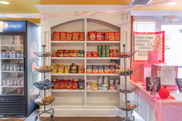 Interior view of a snack and beverage area with shelves stocked with various chips and canned goods. To the left, there is a Pepsi-branded refrigerator filled with drinks. To the right, a table covered with a pink tablecloth displays pink gift bags and a sign for a Henry Ford Hospital Game on Cancer fundraiser raffle.
