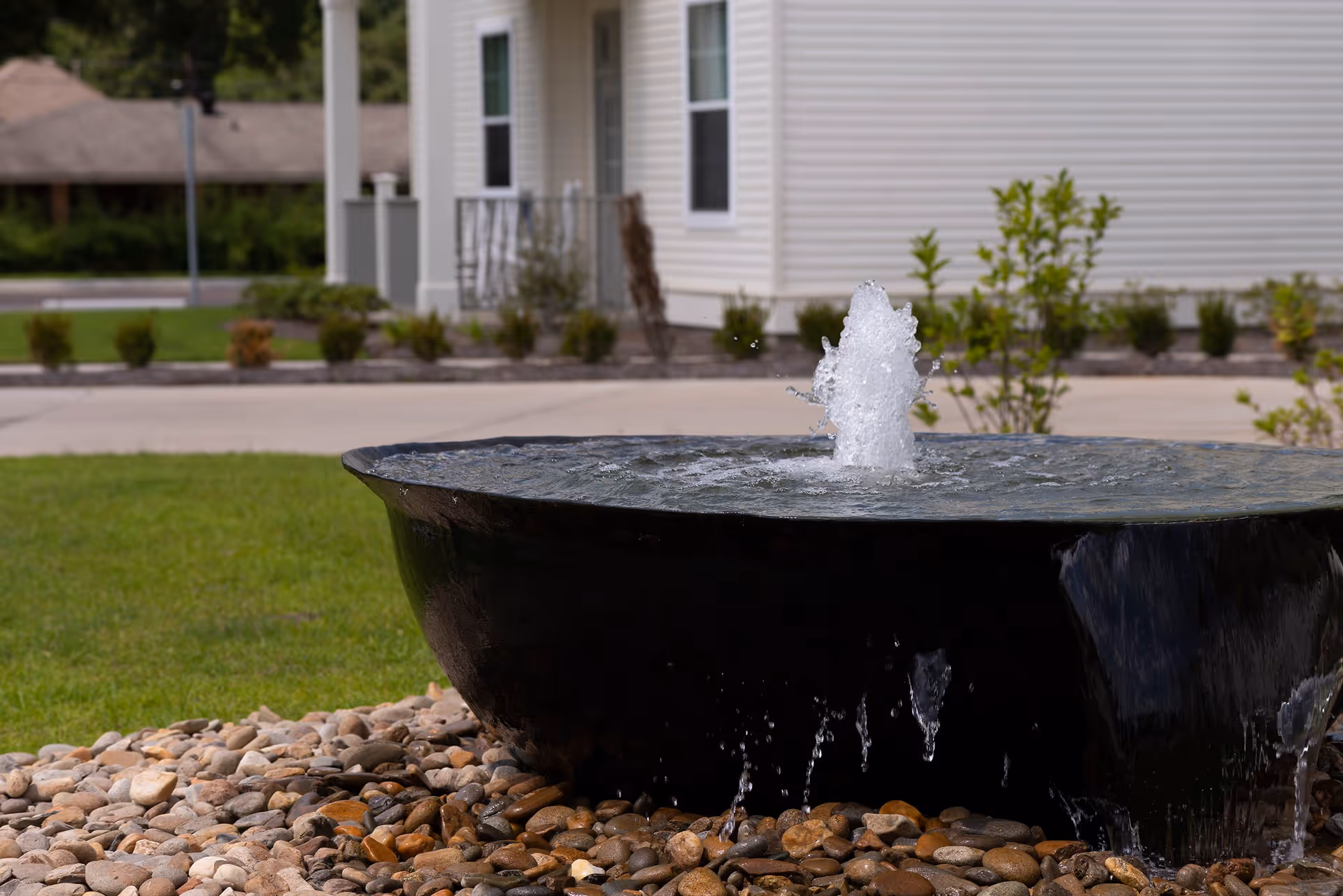 A close-up view of a black water fountain with water bubbling up and flowing over the edges, surrounded by small stones. In the background, there is a green lawn, a paved walkway, and a white building with windows and a porch.