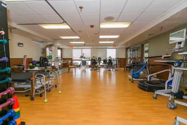 Spacious rehabilitation therapy room with exercise equipment, walkers, and free weights on a wood floor.