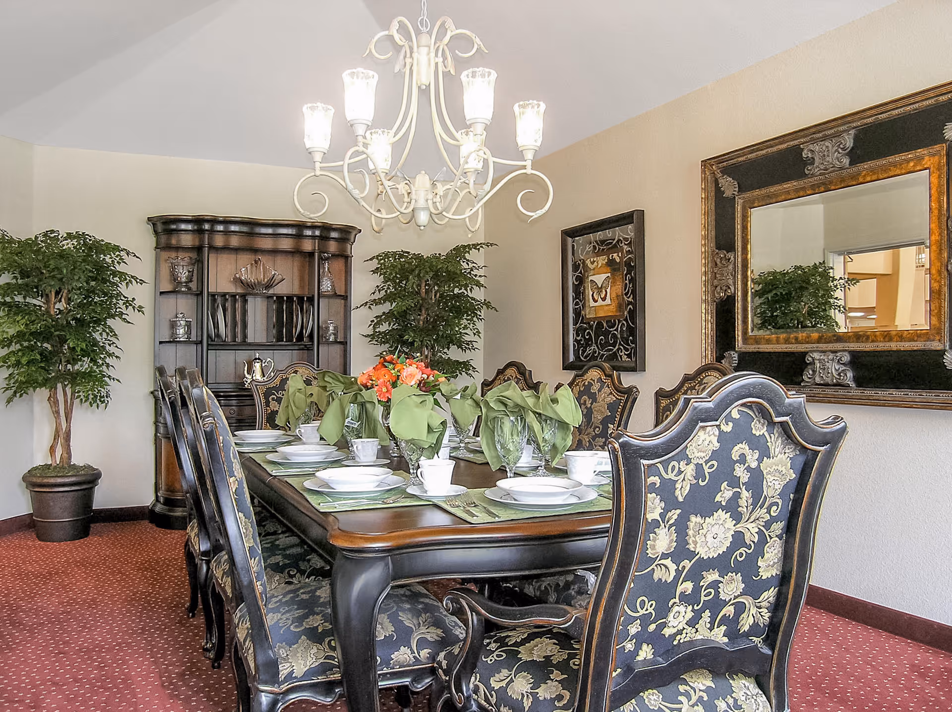 Ornate dining room with a set table, upholstered chairs, chandelier, large mirror, and potted plants.