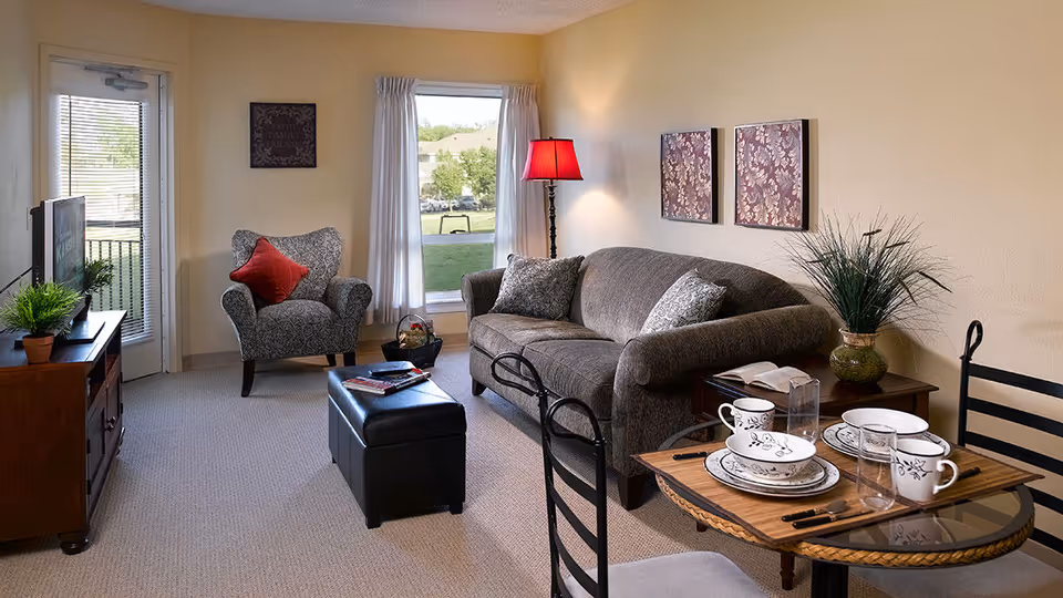 Cozy living room with a sofa and armchair, ottoman, TV cabinet, and a small dining table set beside a window and door.
