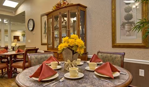 A dining area with a round table set for four, featuring yellow flowers in a vase as a centerpiece, red folded napkins, cups, saucers, and silverware. In the background, there is a wooden china cabinet with glass doors, framed artwork on the walls, and additional tables and chairs.