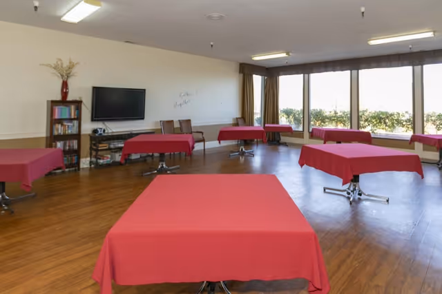 A spacious room with several square tables covered with red tablecloths, wooden flooring, large windows letting in natural light, a bookshelf, a wall-mounted TV, and a few chairs along the wall.