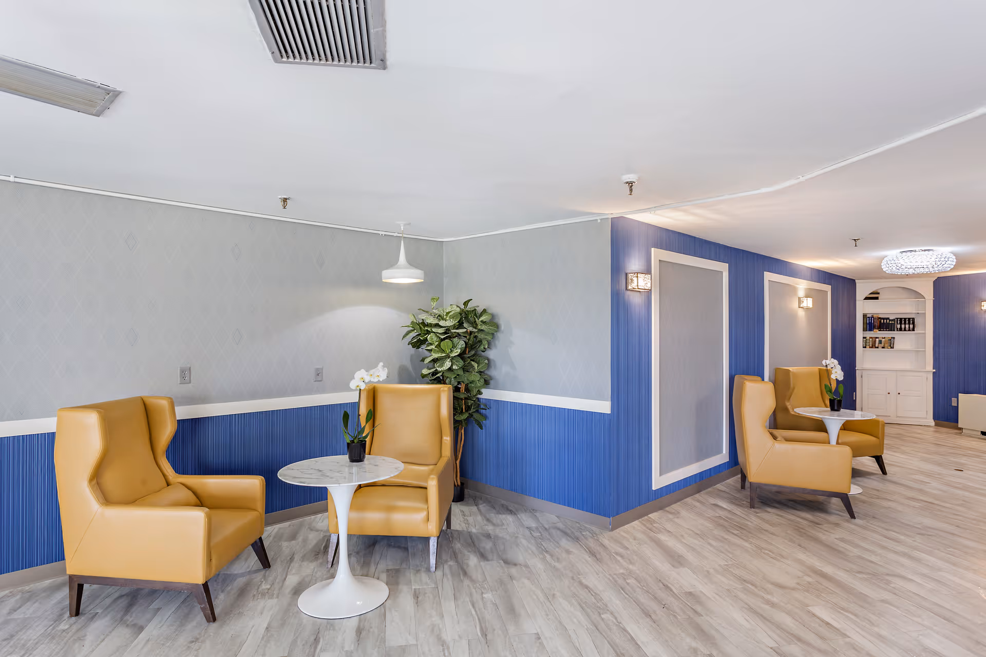 A bright and modern lounge area with light wood flooring, blue and gray walls, and two seating areas each with two mustard yellow armchairs and a small round white table. There are potted plants and decorative lighting fixtures on the walls and ceiling.