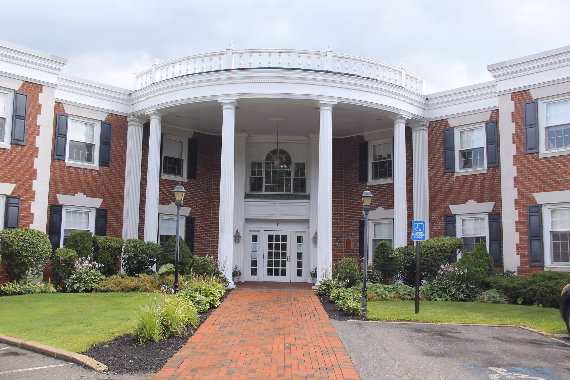 Front entrance of a red-brick building with white columns, a brick walkway, landscaping, and a handicap parking sign.
