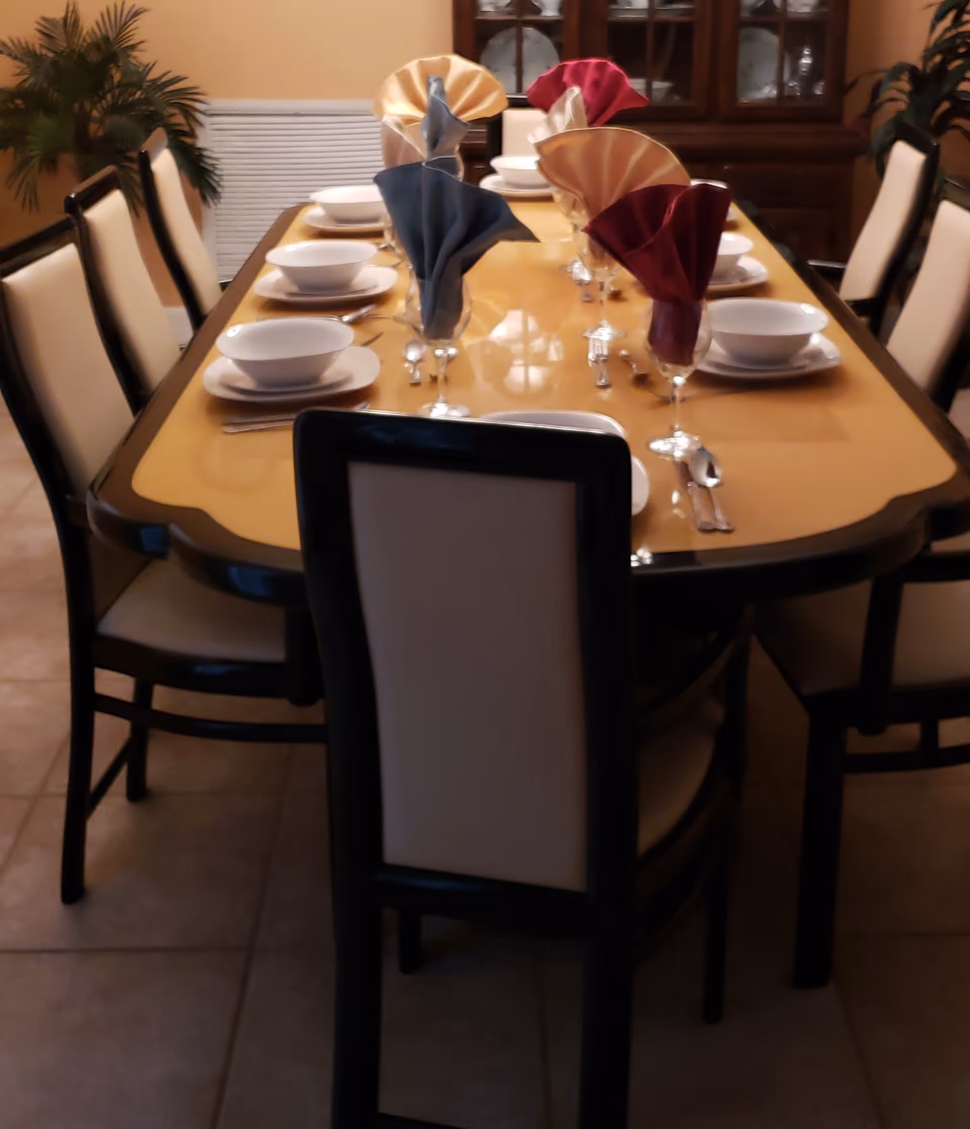 A dining table set for eight people with white bowls, plates, and silverware. Each place setting has a folded cloth napkin in various colors placed in a glass. The table is surrounded by chairs with cream-colored cushions and dark frames. In the background, there is a wooden cabinet with glass doors displaying dishes and plants on either side of the room.