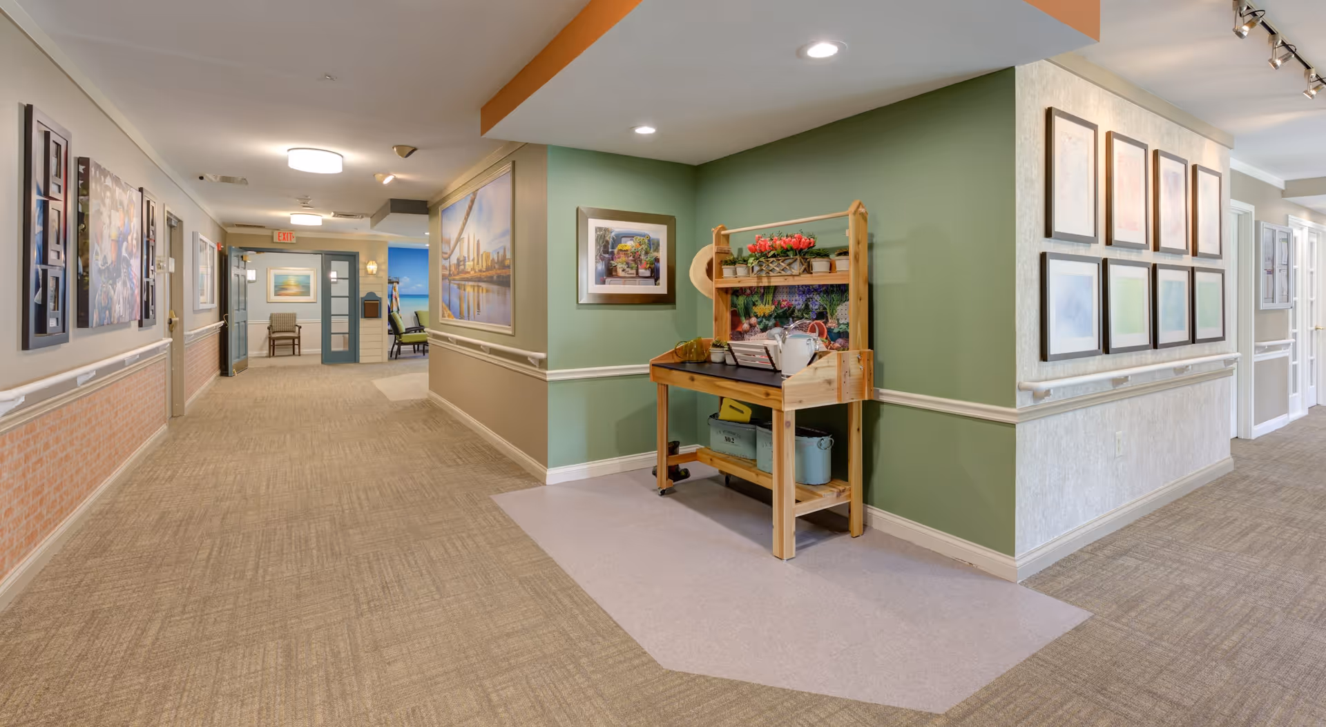 A well-lit hallway in a senior living facility with beige carpet and green and beige walls adorned with framed pictures. A wooden cart with potted plants and gardening tools is placed against one wall. The hallway leads to a room with chairs and more artwork on the walls.