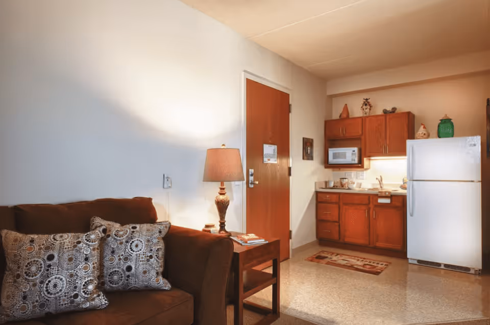 Interior view of a senior living facility unit showing a small kitchen area with wooden cabinets, a microwave, a refrigerator, and a countertop with a sink. Adjacent to the kitchen is a brown couch with patterned pillows, a wooden side table with a lamp, and a closed wooden door in the background.
