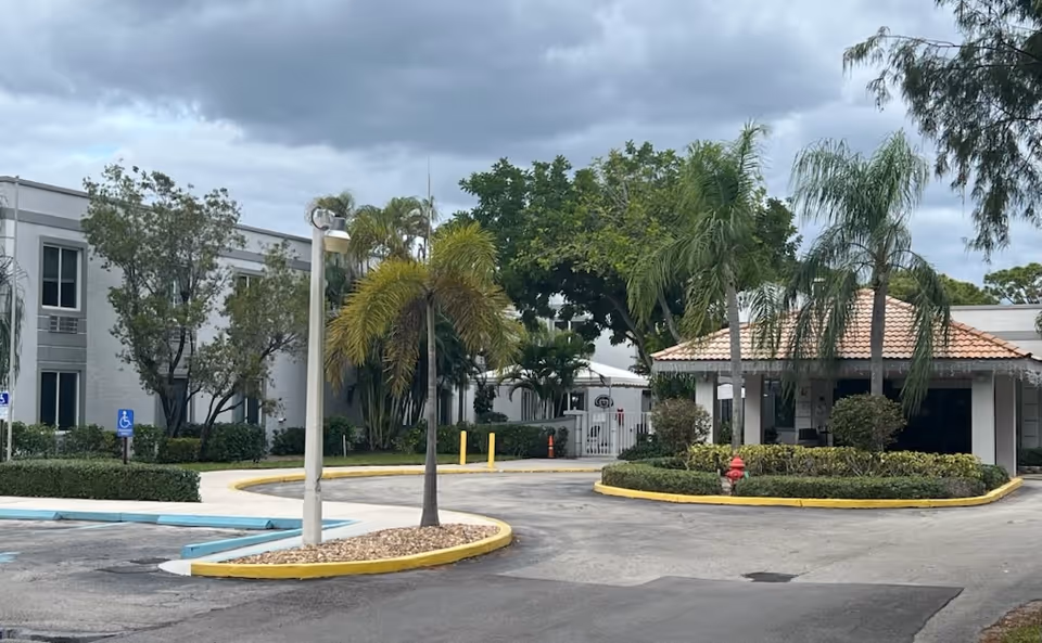 Exterior view of Palm Beach Nursing Center showing a driveway with a circular landscaped island featuring palm trees and bushes. The building has a tiled roof entrance area and is surrounded by more trees and greenery under a cloudy sky.