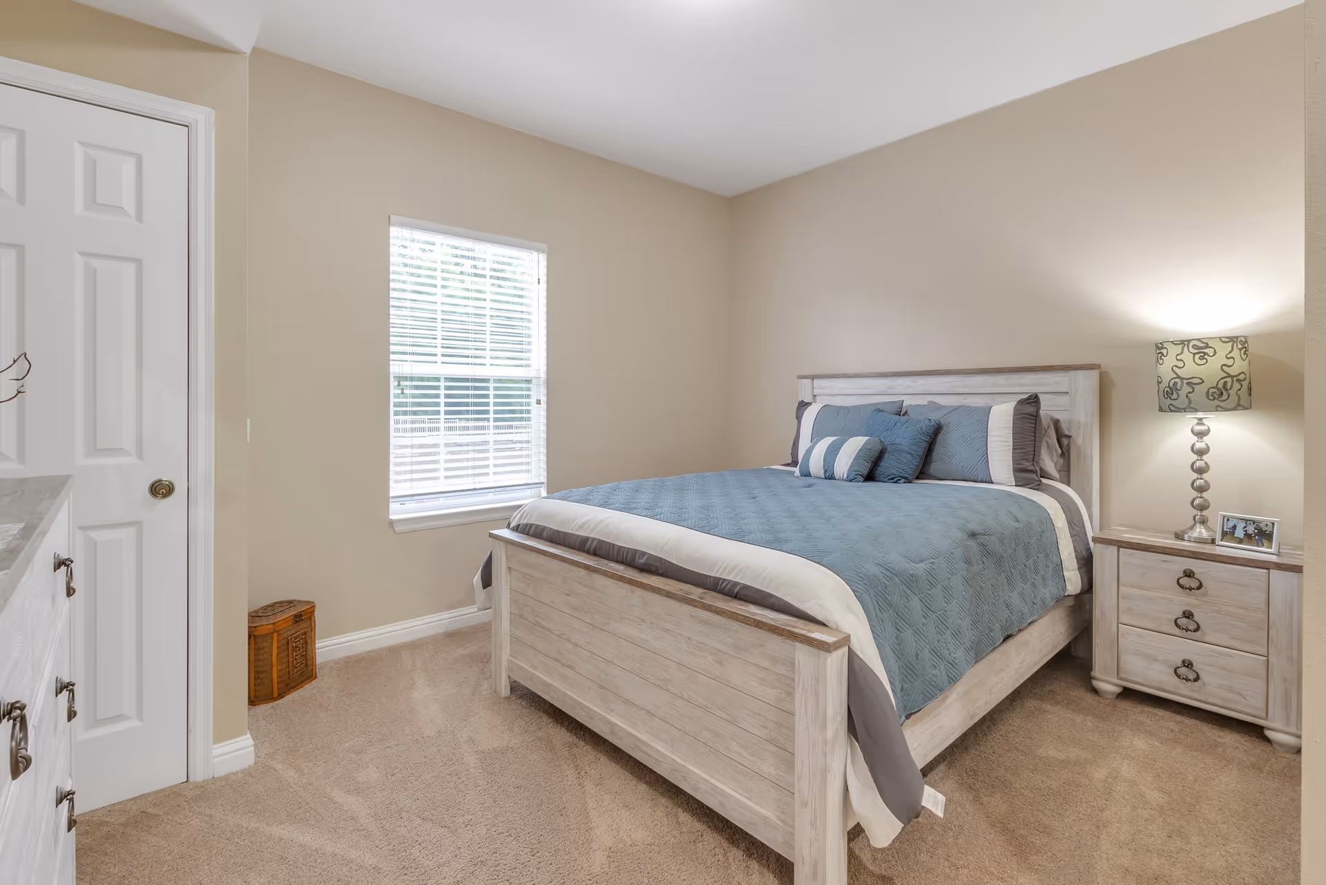 A bedroom with a wooden bed frame and a blue and gray quilted bedspread with matching pillows. There is a wooden nightstand with a lamp and a small framed photo on it. A window with white blinds is on the wall next to the bed, and a white door is visible on the left side of the room. The carpet is beige and the walls are painted a light tan color.