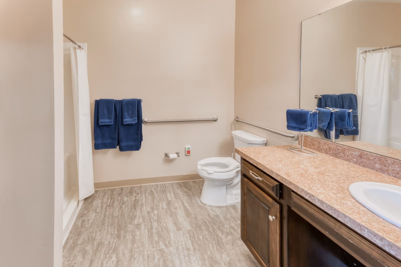 A clean bathroom with a toilet, a shower with a white curtain, a countertop with a sink, and a large mirror. There are blue towels hanging on a towel rack and folded on a stand on the countertop. The walls are beige and the floor has a light wood-like pattern. Safety grab bars are installed near the toilet and shower.