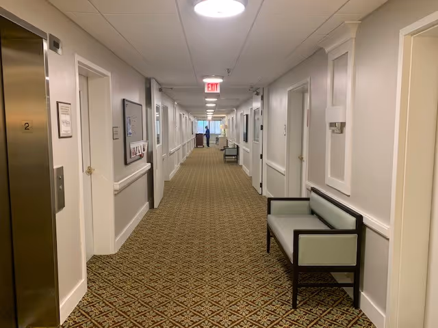 Long carpeted interior hallway with benches, doors, and an elevator leading to an exit sign.