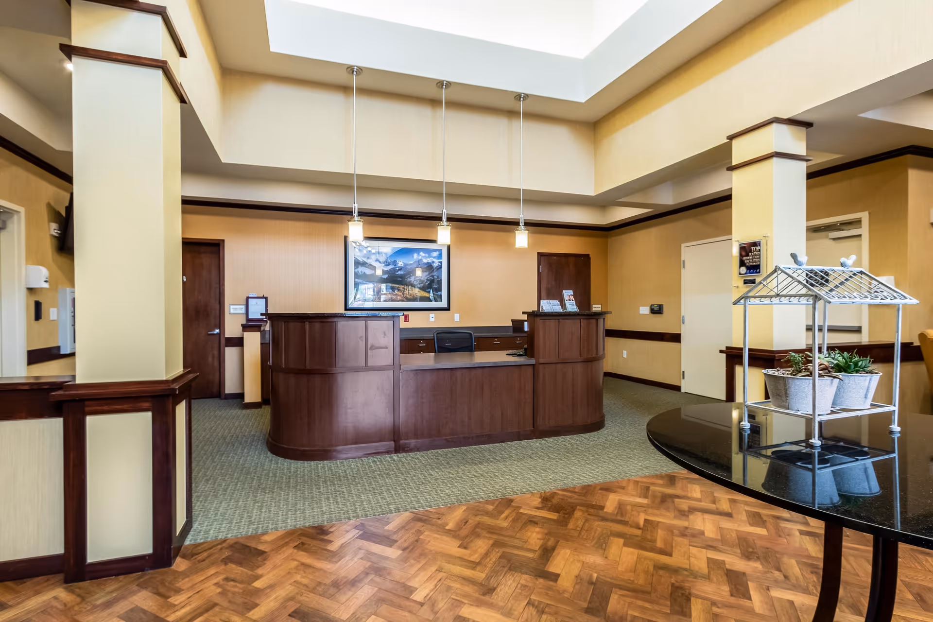 Reception area with a curved wooden desk, three hanging pendant lights, a framed picture on the wall, and a round table with potted plants in a senior living facility.