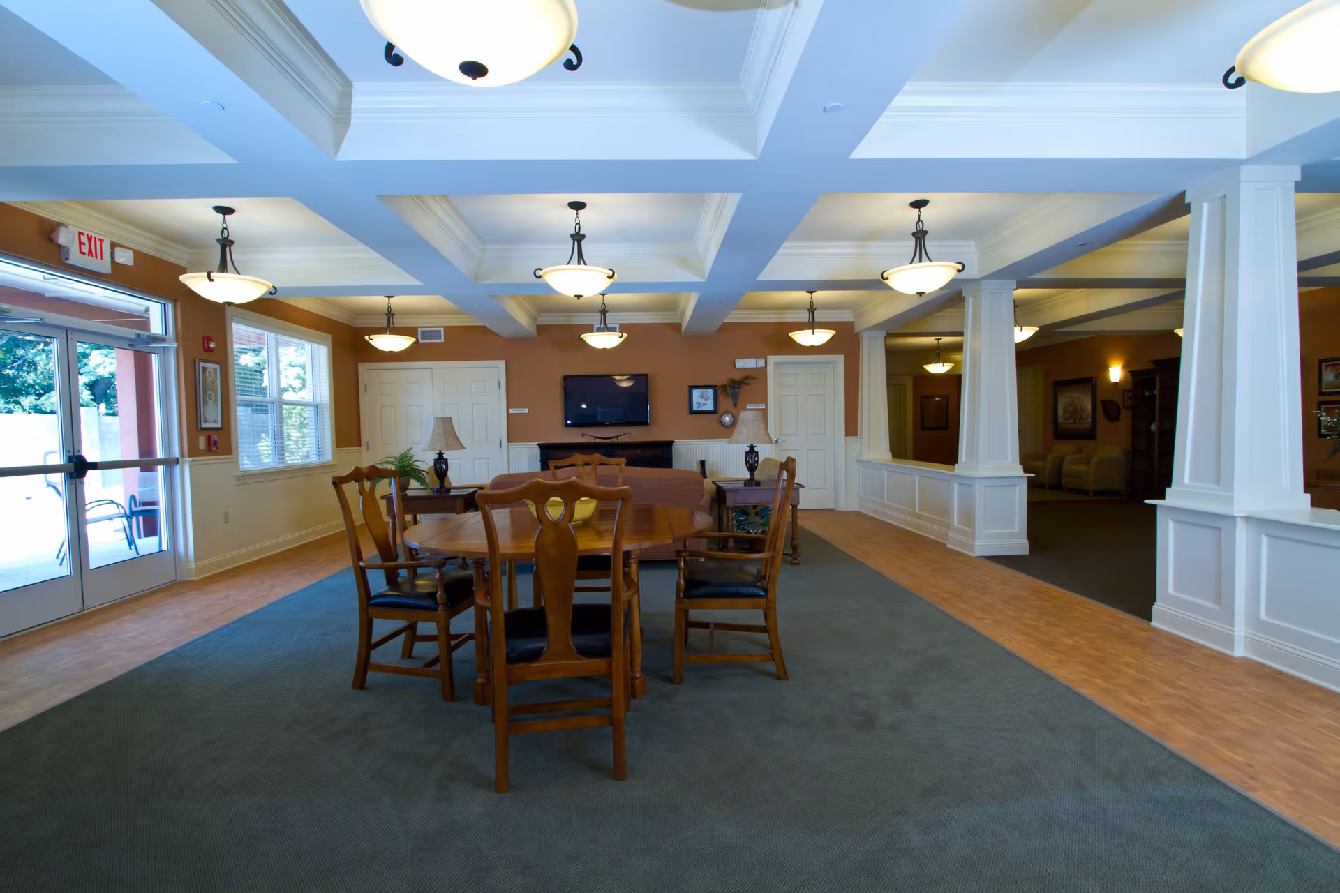 Interior view of a senior living facility common area with a wooden table and chairs in the center, a sofa behind the table, two side tables with lamps, a wall-mounted TV, and multiple ceiling lights. There is a glass door exit on the left and a hallway with seating visible on the right.