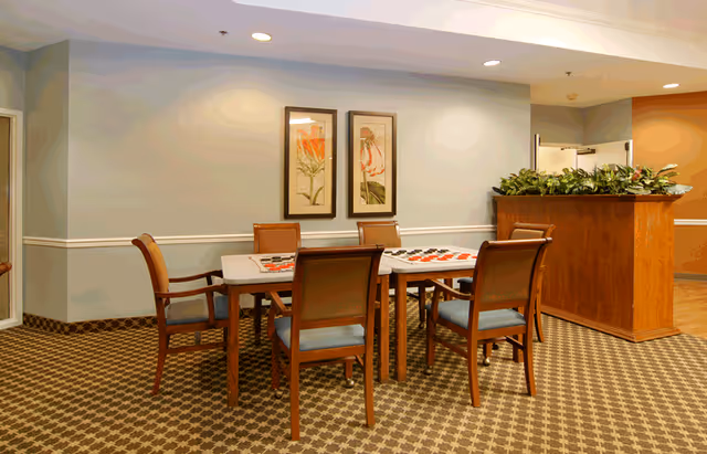 Carpeted dining/activity room with a rectangular wooden table surrounded by six chairs, framed floral artwork on the wall, and a plant-topped wooden partition.