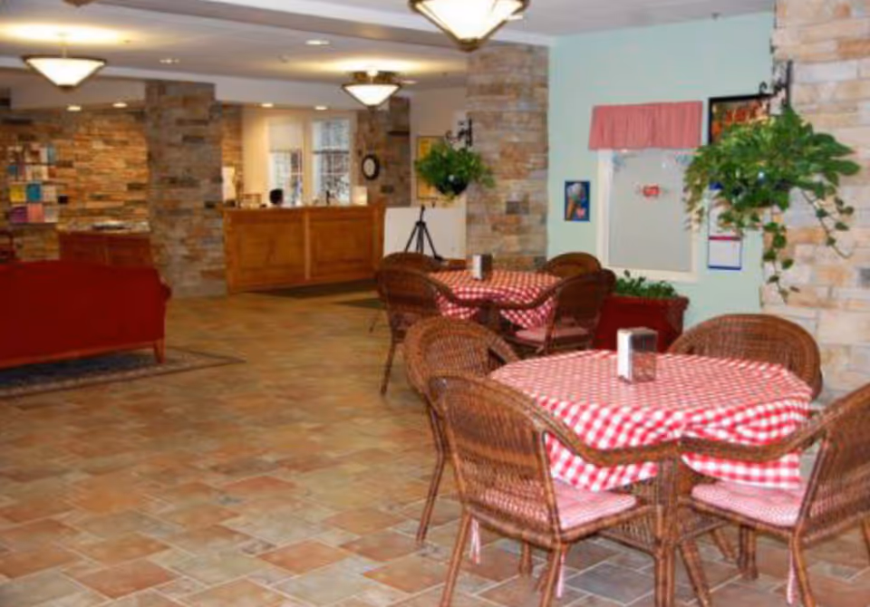 Interior view of a senior living facility lounge area with wicker chairs and tables covered with red and white checkered tablecloths. The room has stone walls, hanging plants, and warm lighting fixtures. A wooden reception desk is visible in the background along with a red couch on the left side.