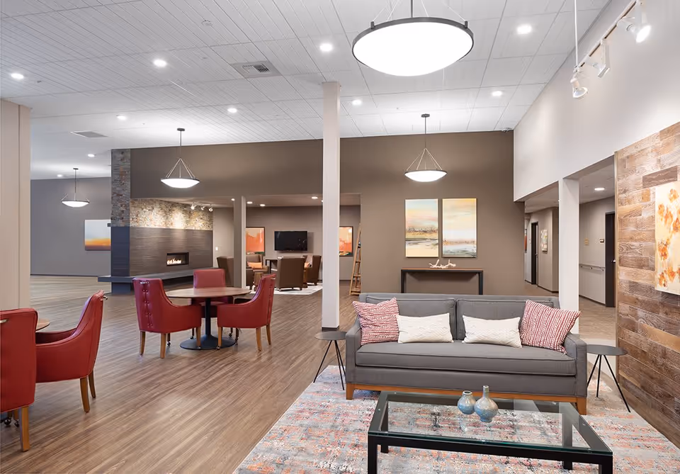 A spacious and modern living room area in a senior living facility featuring a gray sofa with patterned pillows, a glass coffee table on a colorful rug, red armchairs around a round table, a fireplace with a stone accent wall, and contemporary pendant lighting. The walls are painted in neutral tones with decorative artwork and wood paneling.