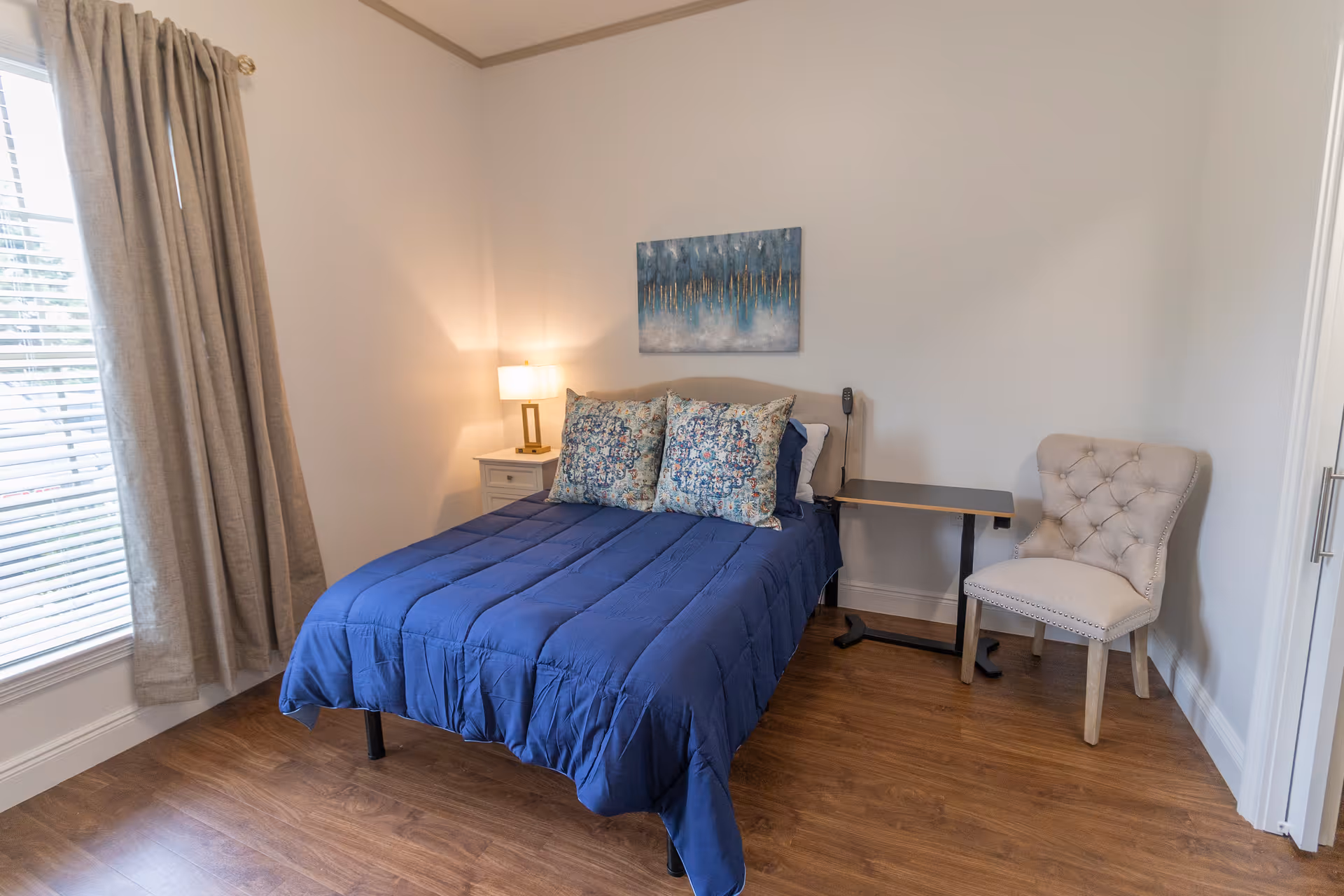 A bedroom with a bed covered in a blue comforter and two patterned pillows. There is a nightstand with a lamp on the left side of the bed, a small adjustable table on the right side, and a beige upholstered chair next to the table. A window with beige curtains is on the left wall, and a blue and gold abstract painting hangs above the bed. The floor is wooden.
