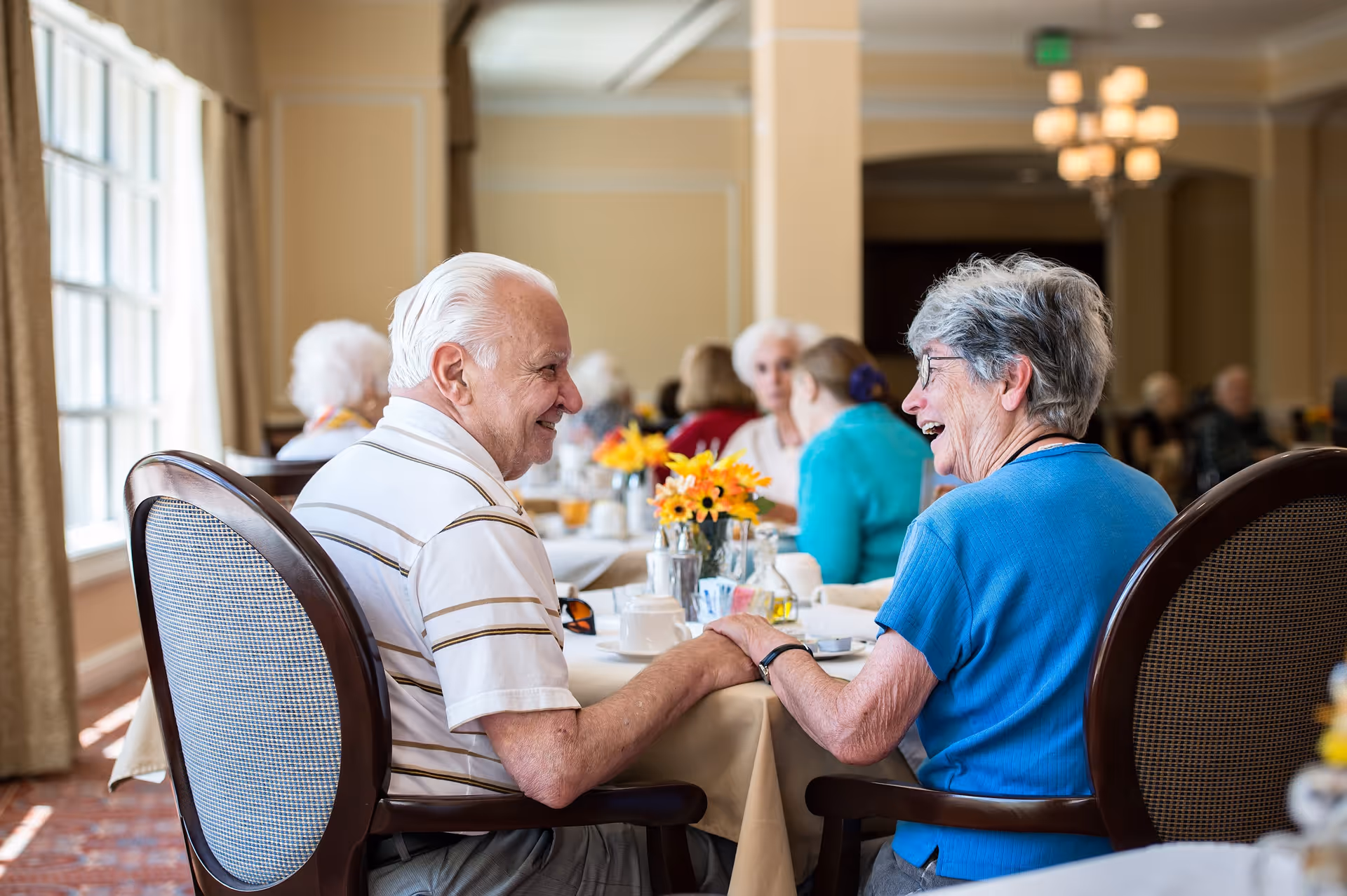 An elderly man and woman sitting at a dining table in a retirement community, holding hands and smiling at each other. Other seniors are seated at tables in the background, with flowers and dining items on the tables in a well-lit room with large windows.