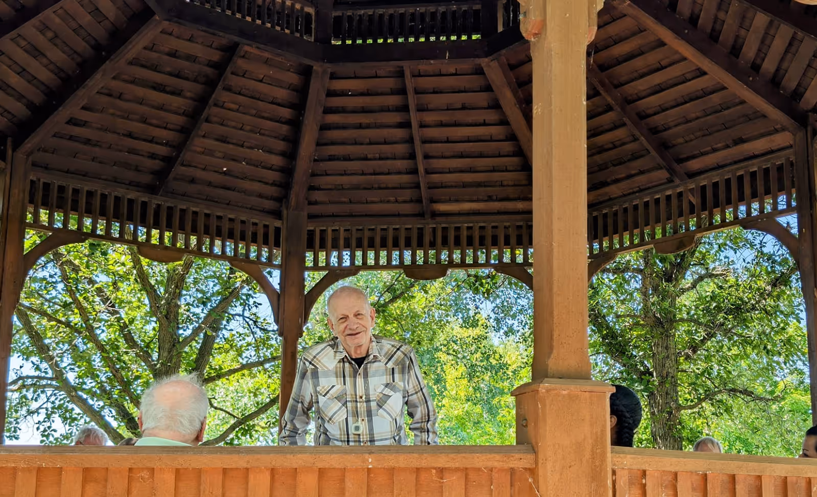 A man stands inside a wooden gazebo with trees visible through the open sides and other people partially visible at the railing.
