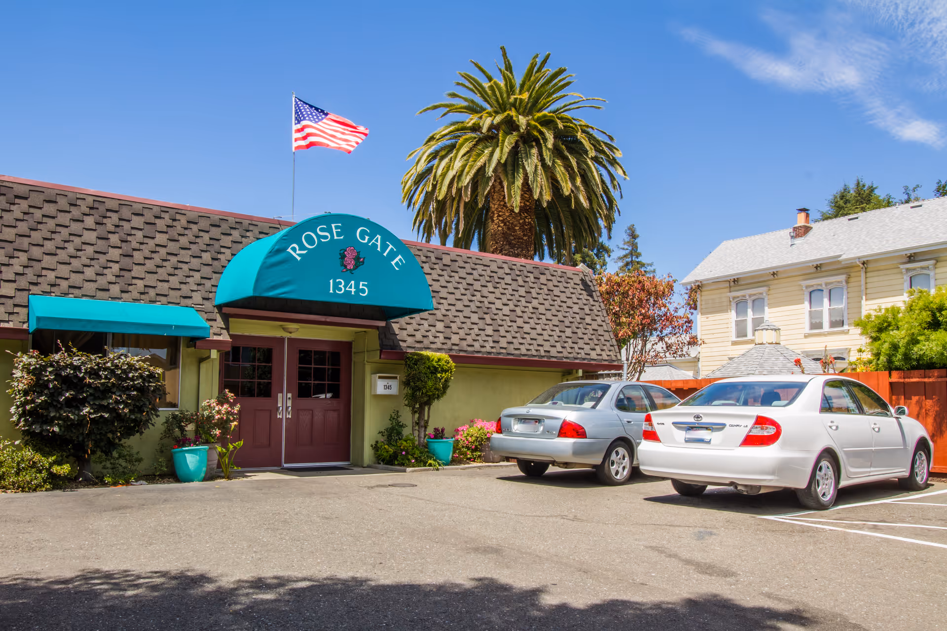 Front entrance of a building with a teal awning reading "ROSE GATE 1345", an American flag and palm tree overhead, and cars parked in front.