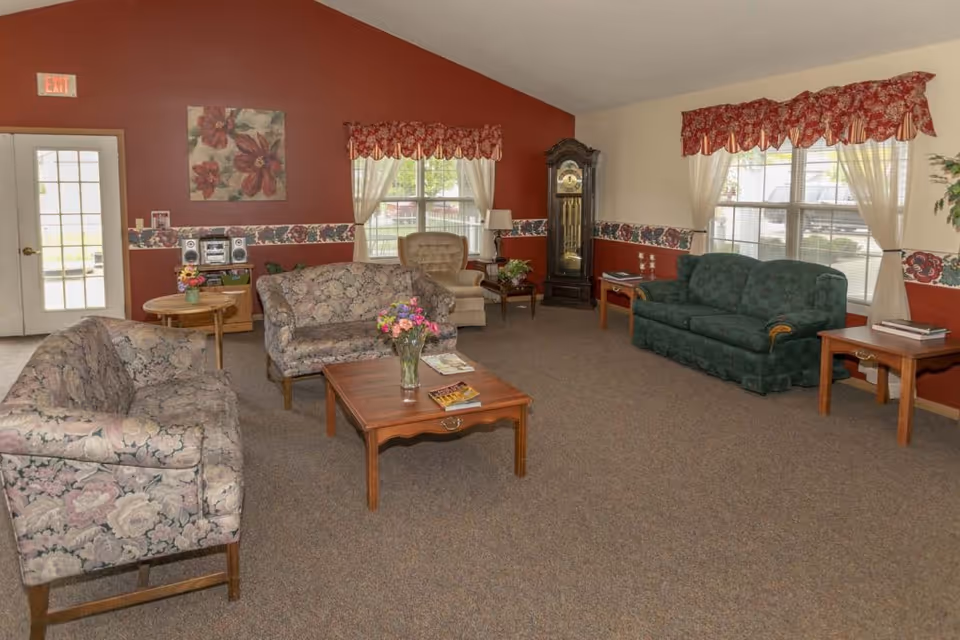 A cozy living room in an assisted living facility with floral patterned sofas, a green couch, a beige armchair, wooden coffee and side tables, a grandfather clock, and windows with red floral valances and white curtains. The walls are painted red and beige with a floral border, and there is a painting of flowers on the wall.