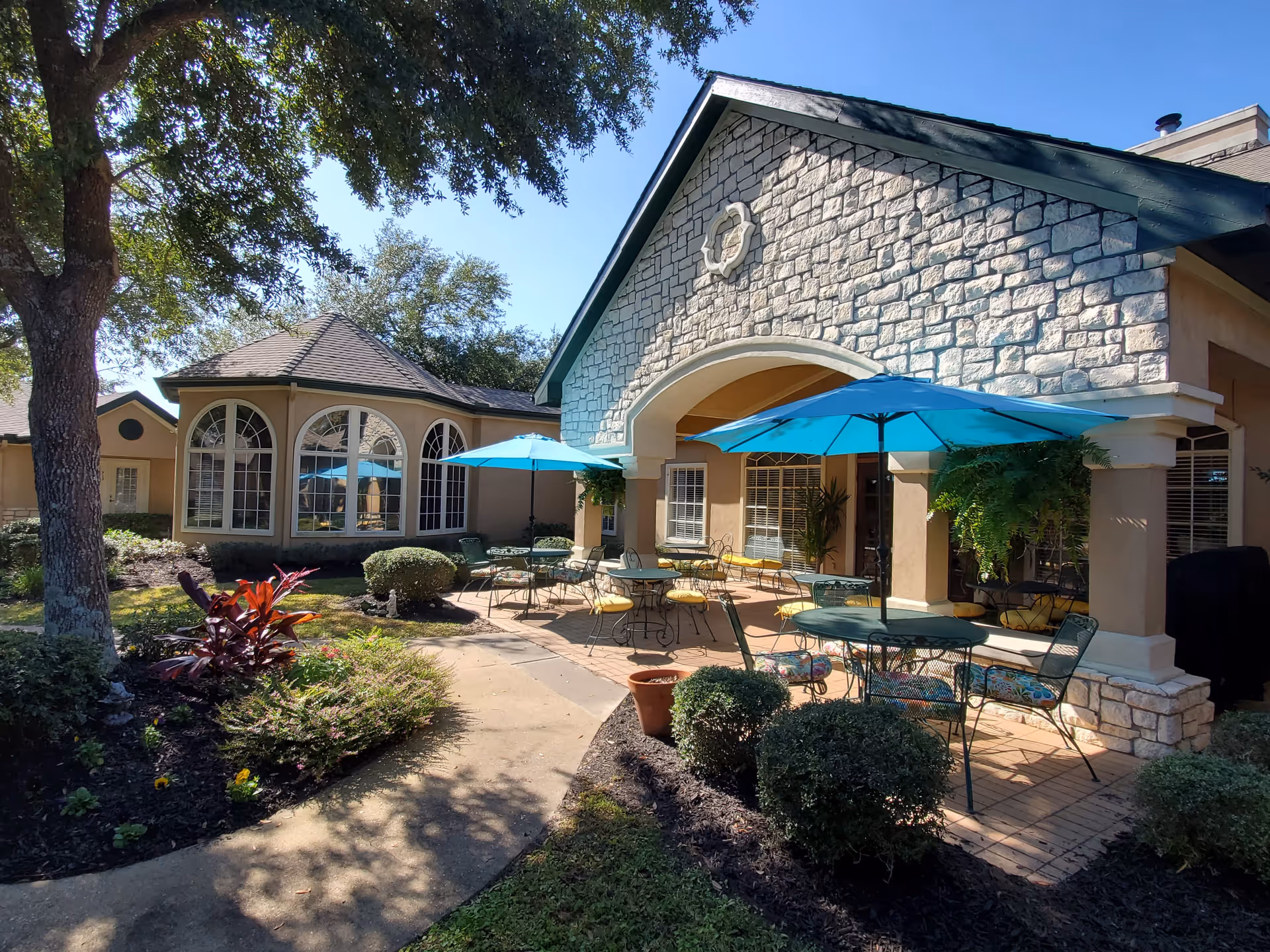 Outdoor patio area at Carriage Inn Huntsville featuring several round tables with blue umbrellas and metal chairs with cushions. The patio is adjacent to a building with stone and stucco exterior walls, large arched windows, and surrounded by landscaped bushes and trees under a clear blue sky.