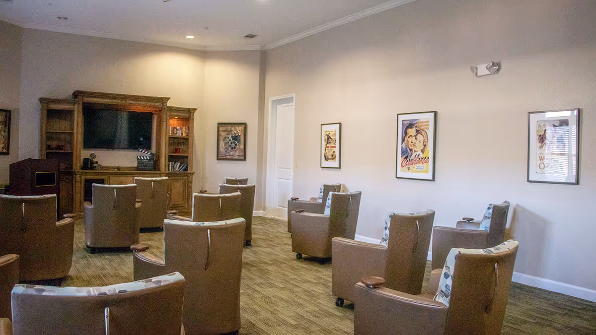 A cozy common area with multiple brown armchairs arranged in rows facing a large wooden entertainment center with a flat-screen TV. The walls are decorated with framed vintage movie posters, and the room has soft lighting and carpeted flooring.