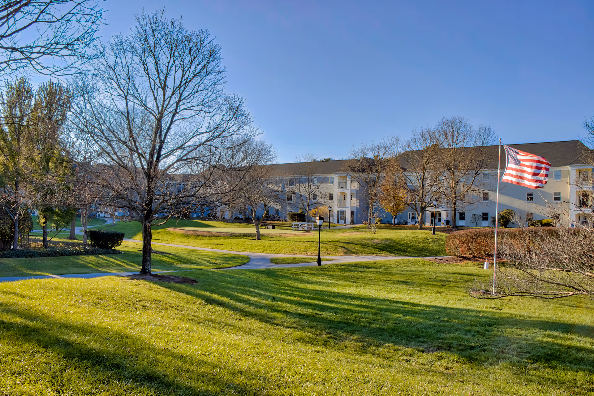 A large grassy outdoor area with a few leafless trees and a paved walkway leading towards a three-story building in the background. An American flag is flying on a flagpole to the right side of the image under a clear blue sky.