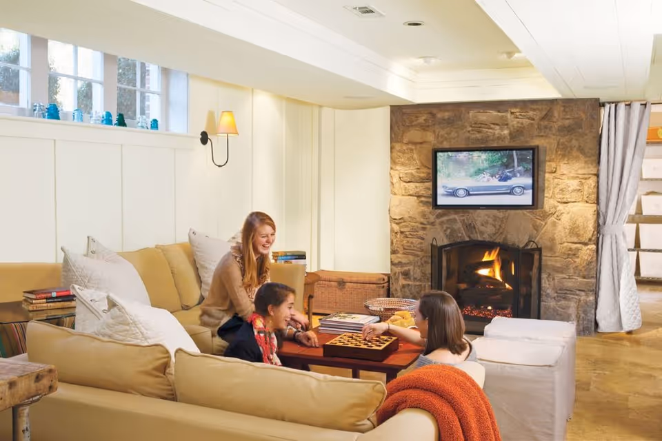 Cozy living room with a stone fireplace and TV, a beige sectional sofa where three people sit around a coffee table playing a board game.
