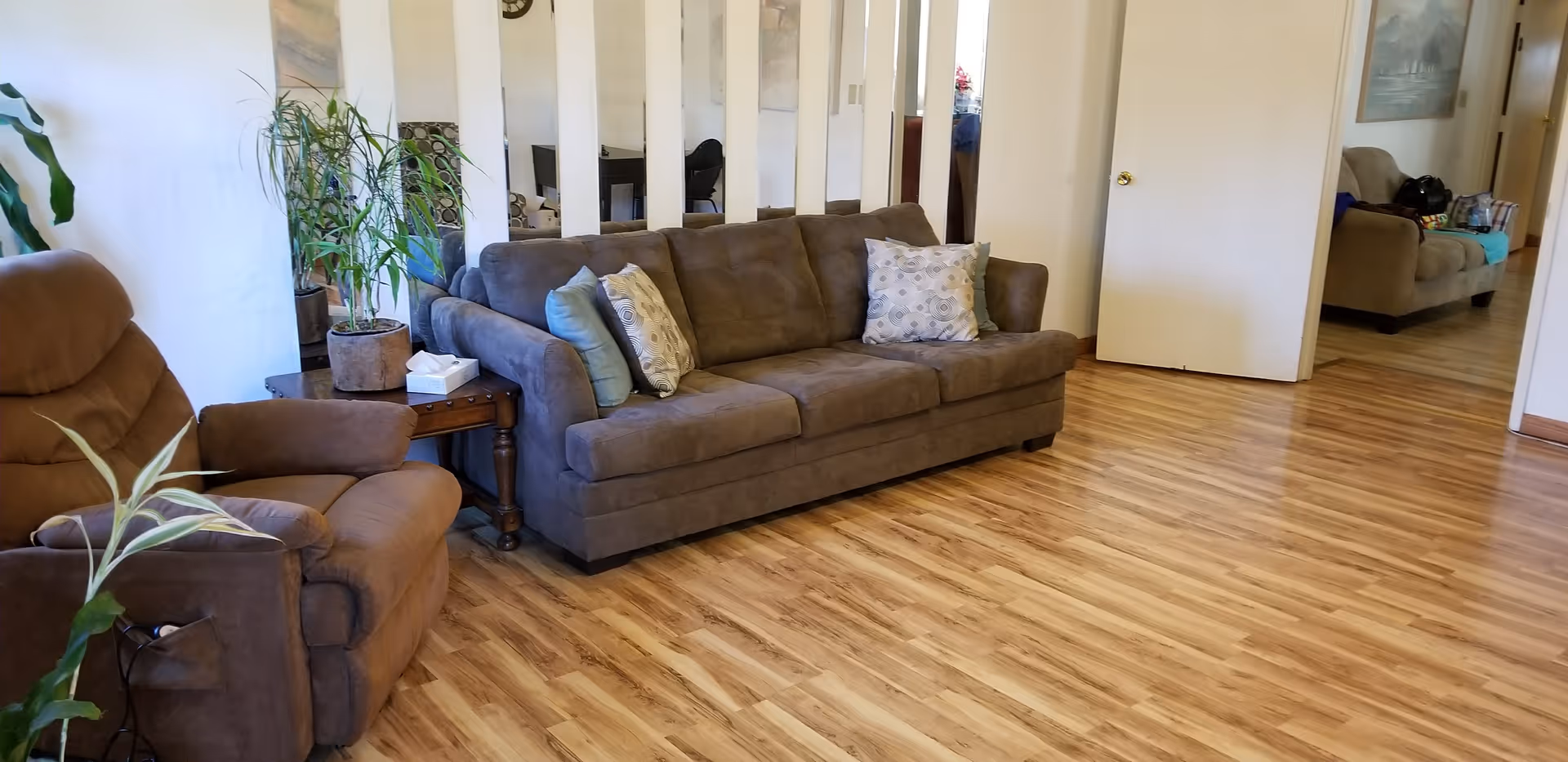 A cozy living room with a brown sofa and a matching armchair. The sofa has several decorative pillows. There is a wooden side table with a tissue box and a potted plant next to the armchair. The floor is wooden, and the walls are light-colored. A large mirror with vertical panels is mounted on the wall behind the sofa, reflecting parts of the room. Another seating area with a couch and a painting on the wall is visible through an open doorway.