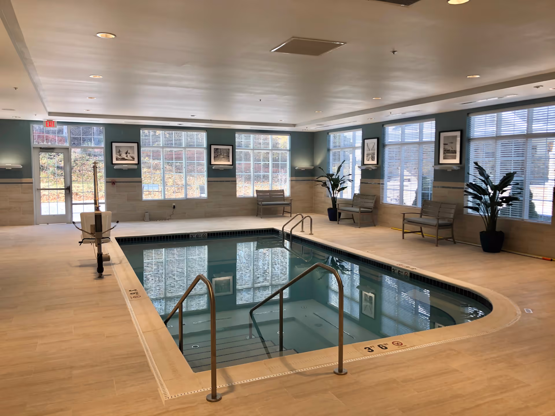 Indoor community swimming pool with handrails, seating benches, large windows, and potted plants.