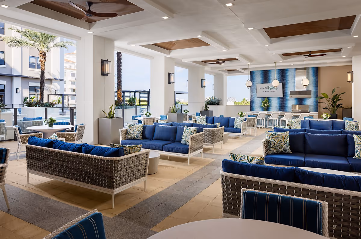 Spacious outdoor seating area at Belmont Village Senior Living Coral Gables featuring multiple wicker sofas with blue cushions and patterned pillows, round tables with chairs, large potted plants, ceiling fans, and a wall-mounted TV above a modern grill setup with a blue textured accent wall.