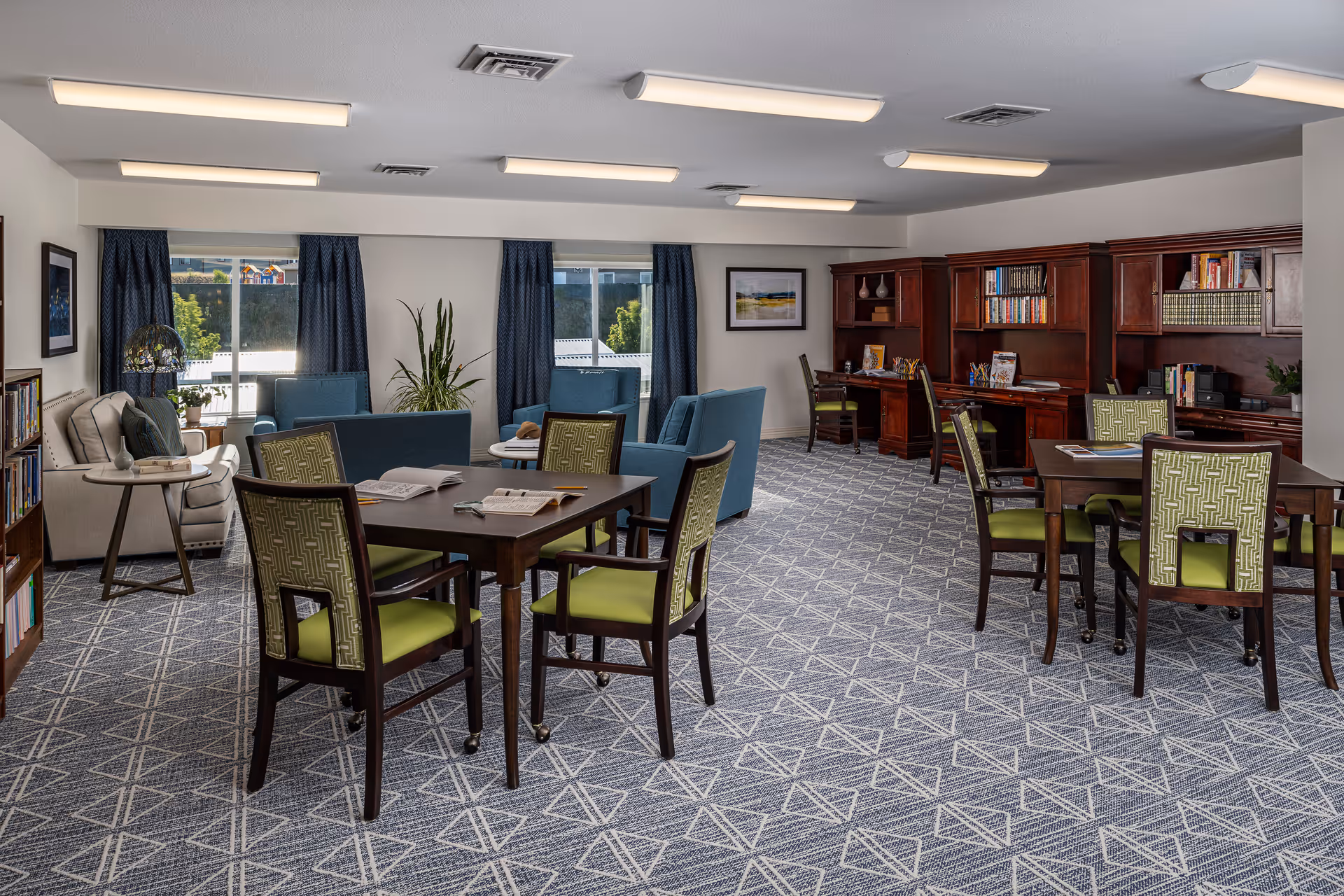 A spacious senior living common area with patterned carpet, several tables with green cushioned chairs, blue armchairs, a beige sofa, wooden bookshelves filled with books, and large windows with blue curtains letting in natural light.