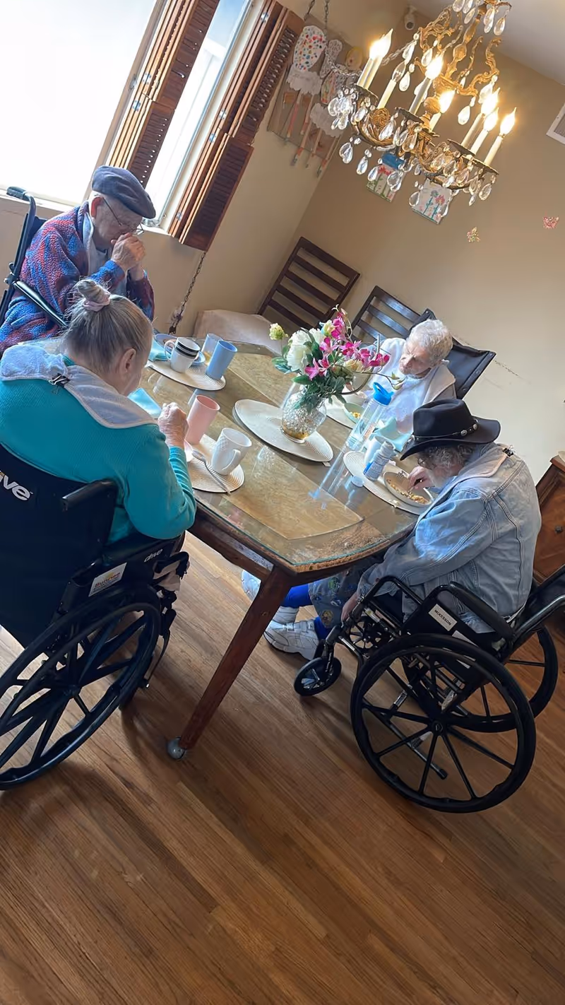 Four elderly individuals, three in wheelchairs, sitting around a dining table in a well-lit room with wooden floors. They appear to be eating or drinking. The table has a vase with flowers in the center, and there is a chandelier hanging above. The room has a window with wooden shutters and some decorations on the wall.