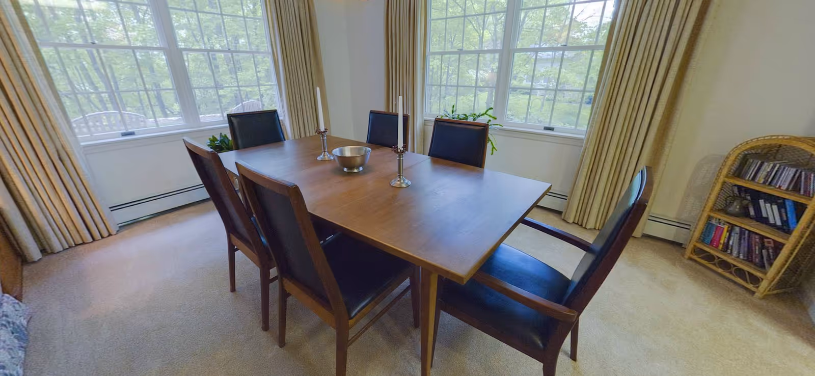 A dining room with a wooden table surrounded by six chairs with dark cushions. On the table are two silver candlesticks with white candles and a silver bowl. Large windows with beige curtains let in natural light, and a small bookshelf with books and CDs is visible in the corner.