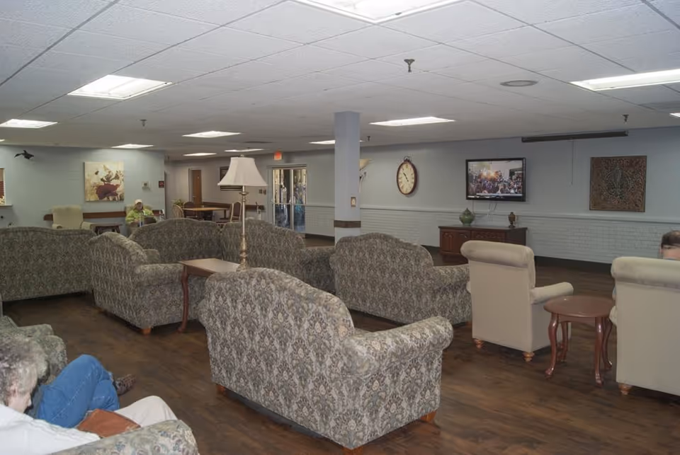 A spacious senior living facility common area with multiple patterned sofas and armchairs arranged around small tables. The room has wood flooring, a white ceiling with fluorescent lights, and light-colored walls decorated with a clock, paintings, and a mounted TV showing a crowd scene. A floor lamp stands near the sofas, and a few seniors are seated in the room.