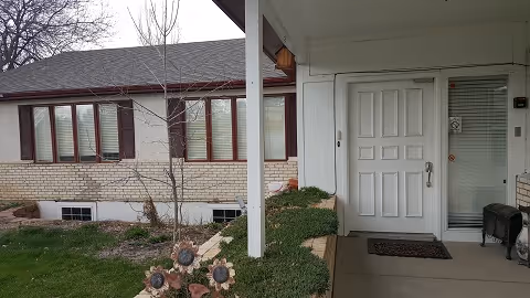 Front entrance of a residential assisted living facility showing a white door with a glass side panel, a small porch area with a doormat, a black bench, and a garden with decorative flowers and shrubs. The building exterior features brick and siding with several windows.
